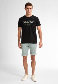 T-shirt nera in cotone con testo bianco che presenta "Petrol Ind." e dettagli grafici, abbinata a pantaloni corti verdi chiari arrotolati e sneakers bianche.