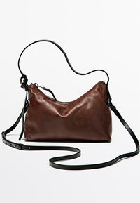 Borsa a mano in pelle marrone con una forma morbida e curvata. Presenta una tracolla nera regolabile e una chiusura superiore con zip. Texture liscia, design minimalista.