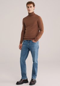 Rjav turtleneck pulover v kombinaciji s svetlo modrimi jeans hlačami in temnimi usnjenimi čevlji. Pulover ima prilega obliko in mehak material.