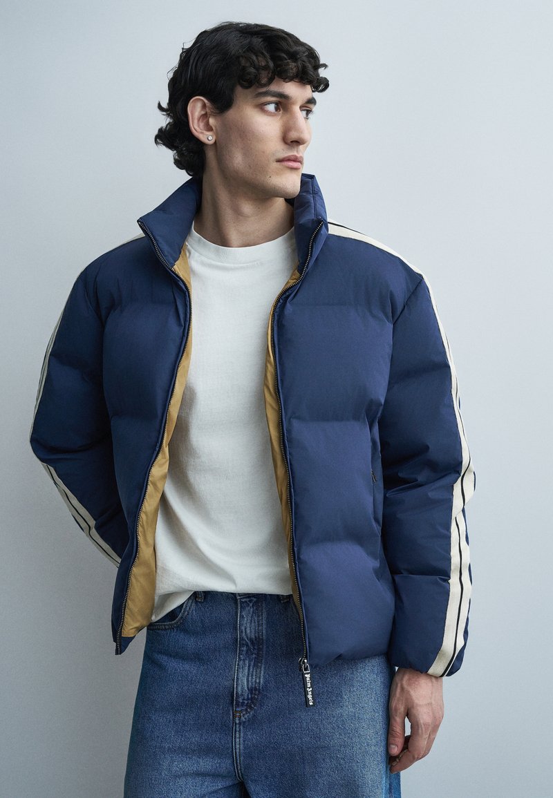 Noor mees kannab valget särki, sinise ja beeži värvi sulejope ning siniseid teksaseid, vaadates vasakule neutraalse tausta ees.