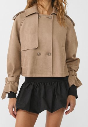 Nainen pukeutuneena lyhyeen beigeen kaksiriviseen mokkatakkkiin, jossa on napilliset olkatoppaukset ja kalvosimet, yhdistettynä mustaan, röyhelöiseen minihameeseen.