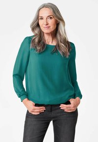 Grüne Chiffonbluse mit langen Ärmeln und rundem Ausschnitt, kombiniert mit dunkelblauen Jeans; glatte Textur mit lockerer Passform und leichtem Glanz.