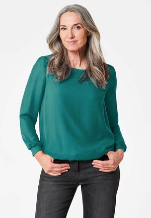 Blusa in chiffon verde con maniche lunghe e scollo rotondo, abbinata a jeans scuri; texture liscia con vestibilità morbida e leggero lucido.