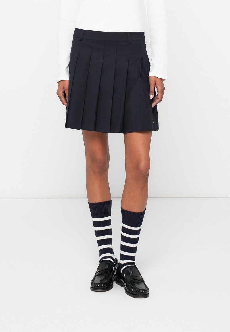 Σκούρο μπλε πλισέ φούστα φτιαγμένη από ύφασμα με λεία υφή. Συνδυάζεται με σκούρο μπλε και λευκές ριγέ καλτσόν μέχρι το γόνατο και μαύρες πατέντες loafers.