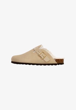 Chausson beige avec un extérieur en daim doux, sangle réglable avec une boucle en or et une semelle en liège texturée. Doublure en polaire confortable visible.