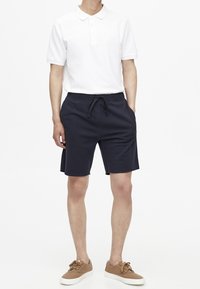 Polo bianco con colletto, abbinato a pantaloni corti blu navy con vita elastica e tasche laterali. Scarpe marroni con lacci completano il look.