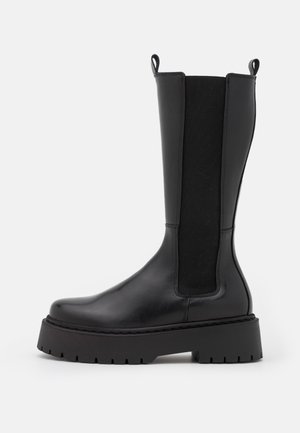 Botte noire en cuir haute jusqu'au genou avec semelle épaisse à crampons, panneau latéral élastique et languettes en haut, sur fond blanc.