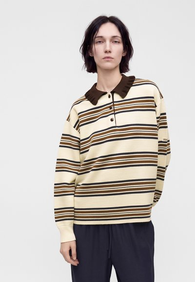 Personne aux cheveux courts et foncés portant une chemise beige à manches longues avec des rayures horizontales marron et noire et un col froncé marron foncé.