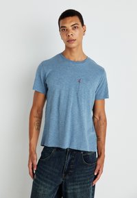 Kék, kerek nyakú póló, puha anyagból készült, kis mellzsebbel és rövid ujjú kivitelben, sötét denim nadrággal párosítva.