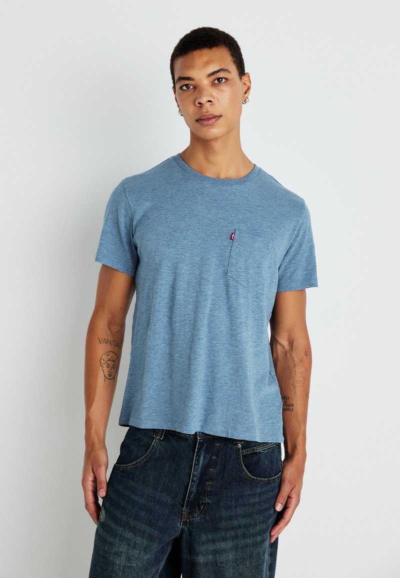 Kék, kerek nyakú póló, puha anyagból készült, kis mellzsebbel és rövid ujjú kivitelben, sötét denim nadrággal párosítva.