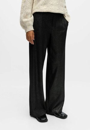 Person wearing wide-leg black shiny pants, black square-toe shoes, and a cream textured knit sweater with hand in pocket.