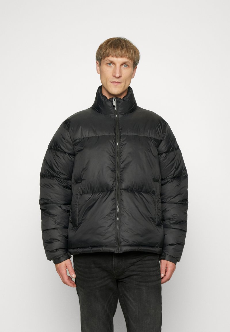 Veste doudoune noire avec un col haut, fermeture éclair et coutures horizontales. Fabriquée en matériau lisse et brillant, avec des poches latérales.