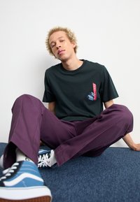 Tricou scurt verde închis cu accent grafic roșu și albastru, purtat cu pantaloni purpurii lejeri și pantofi sport albaștri.