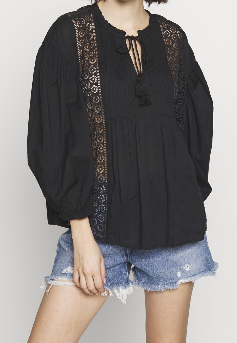 Femme portant un chemisier noir à manches longues avec des panneaux en dentelle et un lien à pompons au niveau du cou, associé à un short en denim bleu déchiré.