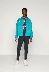 Kvinne iført en teal mønstret jakke over en flerfarget cropped topp, svarte leggings og hvite joggesko, stående mot en ensfarget bakgrunn.