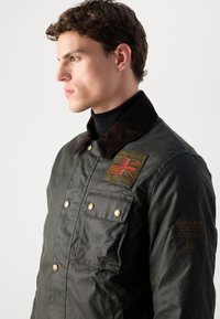 Veste en coton ciré de couleur olive avec un col en velours côtelé marron, des boutons sur le devant, deux poches poitrine et un détail brodé du drapeau britannique.
