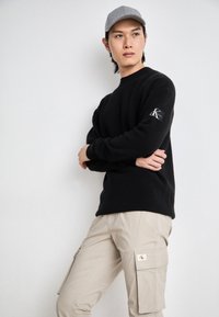 Jersey negro acanalado con un logo en la manga, combinado con pantalones cargo beige que tienen bolsillos laterales y una etiqueta. Gorra gris en la cabeza.