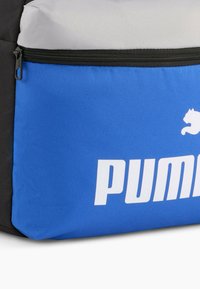 Zaino blu e nero con parte superiore grigia, caratterizzato da un grande logo bianco "PUMA" e una tasca con zip realizzata in tessuto testurizzato.