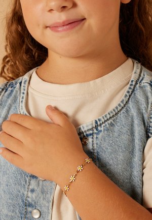 Gouden armband met witte madeliefcharms, gedragen aan een hand. Denim vest, crèmekleurig overhemd zichtbaar. Soepele textuur, delicate ontwerp.