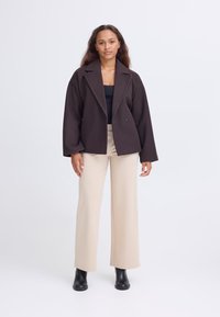 Donna in piedi che indossa un giacketto oversize marrone scuro, top nero, pantaloni beige a gamba larga e stivaletti neri contro uno sfondo neutro.