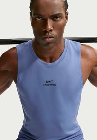 Αθλητικός άνδρας με μπλε αμάνικη μπλούζα Nike Pro κρατά μια μπάρα στους ώμους του κοιτάζοντας συγκεντρωμένα προς τα εμπρός.