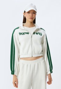 Witte zip-up trainingsjack met groene zijstreep en tekst. Bijpassende sweatpants met elastische tailleband. Katoenmengsel, casual fit.