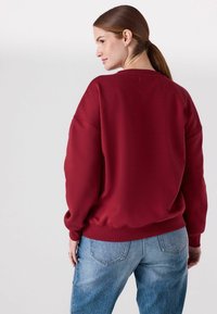 Bordeaux sweatshirt met een losse pasvorm, geribbelde manchetten en zoom. Heeft een ronde halslijn en een effen kleur. Gecombineerd met hoge blauwe jeans.
