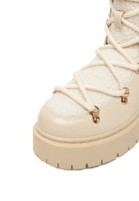 Cizmă platformă bej, cu un superior texturat din fleece, margini din piele și accesorii din metal auriu. Include detalii cu șireturi și o talpă groasă.