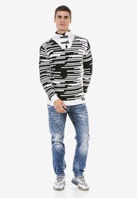 Schwarz-weißer gemusterter Pullover mit hohem Kragen und Knopfdetails, getragen mit blauen, abgetragenen Jeans und grauen Sneakern.