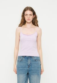 Γυναίκα με κυματιστά καστανά μαλλιά που φοράει χρυσούς κρίκους σκουλαρίκια, ανοιχτό μωβ καμιζόλ με δαντέλα και μπλε τζιν παντελόνι σε λευκό φόντο.