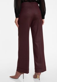 Vrouw in bordeauxrode gestreepte wijde broek en beige hoge enkellaarsjes, gezien vanaf de achterkant, staand op een witte vloer.