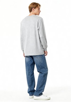 Junger Mann steht seitlich, trägt ein hellgraues Langarmhemd, lockere blaue Jeans und weiße Turnschuhe vor weißem Hintergrund.