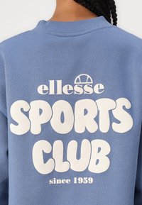Sweatshirt bleu clair avec des lettres blanches en gras qui disent "ellesse SPORTS CLUB" avec le texte "depuis 1959" en dessous. Design classique à col ras du cou.