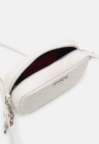 Hvid læder crossbody taske med præget logo-mønster, der har lynlås-lukning og hardware i sølv-tone. Indvendigt er den foret med rødt stof.