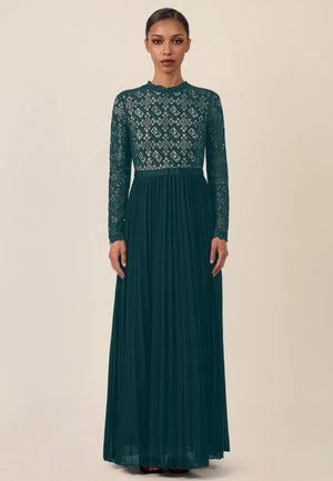 Frau in einem langen dunkelgrünen Kleid mit Spitzenmuster am Oberteil und an den Ärmeln, stehend vor einem schlichten beigen Hintergrund.