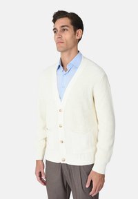 Cardigan bianco lavorato a maglia con scollo a V, bottoni sul davanti, due tasche frontali e polsini e orlo a coste. Indossato sopra una camicia azzurro chiaro.