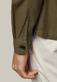 Manga de chaqueta verde oliva con detalle de botón y ajustador en el puño; presenta una textura lisa y acentos de costura cerca del dobladillo.