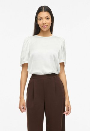 Mujer con cabello largo y oscuro, vestida con una blusa blanca de mangas abullonadas metida dentro de pantalones marrón oscuro de talle alto, frente a un fondo liso.