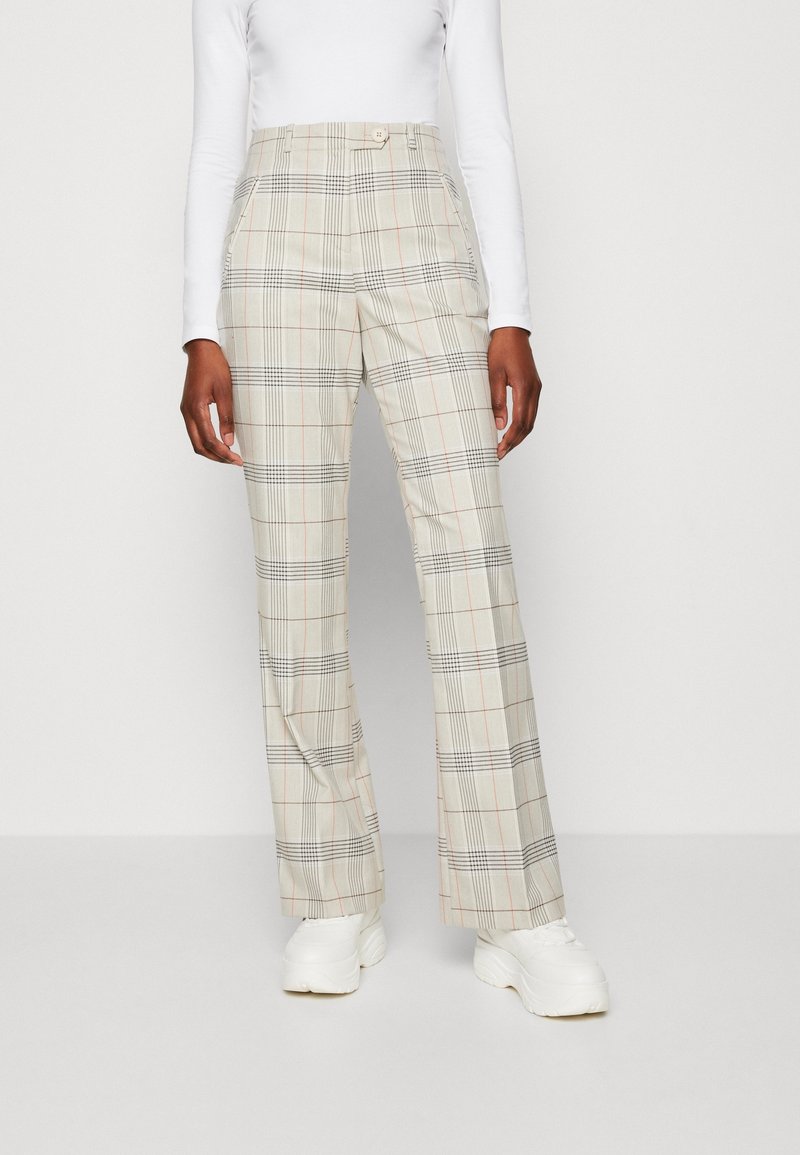 Personne portant un pantalon beige à carreaux taille haute et jambes larges ainsi que des baskets blanches, debout devant un fond blanc uni.