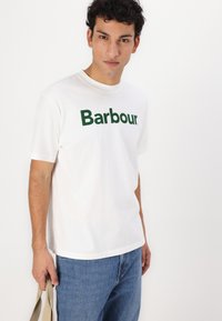 Bílý bavlněný tričko s zeleným logem "Barbour". Krátké rukávy, kulatý výstřih, volný střih, spárované s modrými džínami.