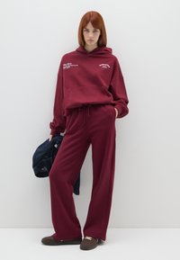 Sudadera burdeos con capucha y cordón, con texto blanco. Pantalones de pierna ancha burdeos a juego, con una chaqueta de mezclilla colgada de un brazo.