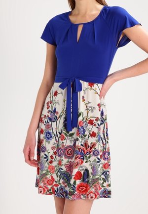 Femme portant une robe à manches courtes bleue avec une jupe à motif floral et une ceinture à la taille, debout avec une main sur la hanche.
