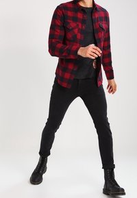 Czerwona i czarna flanelowa koszula w kratę z dwoma przednimi kieszeniami, noszona na czarnej koszulce, w połączeniu z czarnymi spodniami i sznurowanymi butami.