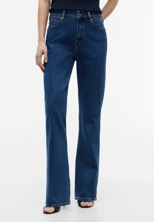 Kvinde iført mørkeblå straight-leg jeans med knap og lynlås, kombineret med sorte åbne hæle og en mørk top.
