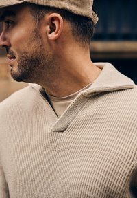 Beige getextureerde breipullover met een brede kraag en een klein tabje bij de halslijn, gedragen over een beige binnenhemd.