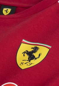 Sarkans kokvilnas T-krekls ar dzelteno Ferrari logo, kurā attēlots melns lecošs zirgs un "SF" teksts, akcentēts ar zaļām un baltām svītrām.