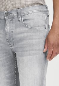 Nahaufnahme von hellgrauen, verwaschenen Jeans mit Vordertasche und einer Hand, die neben dem Oberschenkel liegt, getragen mit einem karierten Hemd, das in der Hose steckt.