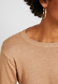 Beige Pullover mit Rundhalsausschnitt, der mit kleinen, hellen Perlen übersäht ist. Eine goldene Creole ist sichtbar.