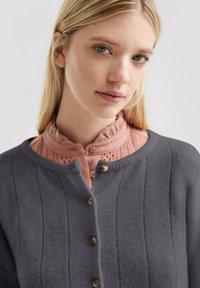Grijze gebreide cardigan met verticale strepen, met een ronde halslijn en vijf bruine knopen, gelaagd over een roze gestructureerde blouse.