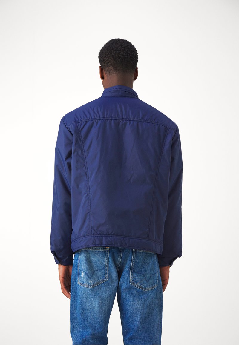 Chaqueta azul marino con cuello alto, mangas largas y un acabado de textura suave, llevada sobre jeans de un azul claro.